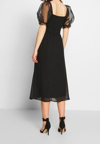 Schwarzes Midikleid mit transparenten, gepufften Ärmeln und einem gerafften Oberteil. Der Rock ist glatt und fließend, kombiniert mit schwarzen High Heels.