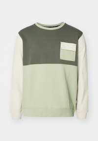 Sudadera color bloque verde y crema con mangas largas, cuello redondo y un bolsillo en el pecho. Fabricada con suave material de algodón.