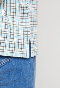 Niebiesko-biała kraciasta koszula z czarnymi liniami tworzącymi siatkę, z bocznym rozcięciem. Noszona z niebieskimi dżinsami, podkreślająca casualowy styl.