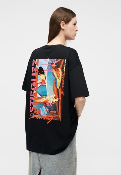 Γυναίκα με καστανά μαλλιά που φοράει μια μαύρη φαρδιά μπλούζα με πολύχρωμη παραθαλάσσια σκηνή και κείμενο στην πλάτη, σε συνδυασμό με μια ανοιχτόχρωμη τζιν φούστα.