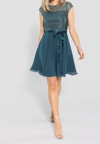 Robe teal avec un corsage à paillettes et une jupe fluide. Dispose d'une taille en satin et d'un nœud de ruban. Texture lisse avec des manches courtes.