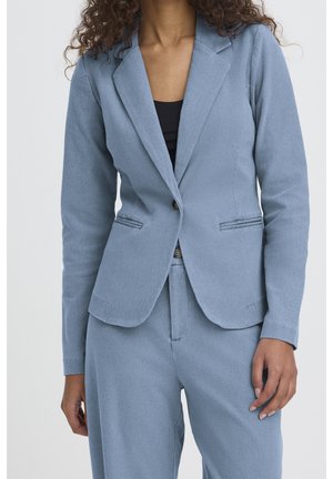 Vrouw draagt een lichtblauwe, op maat gemaakte blazer en bijpassende broek met een zwart topje dat eronder zichtbaar is.