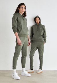 Olivgrüne Hoodies und Sweatpants aus weichem Stoff mit Kängurutaschen. Weiße Sneaker mit strukturierten Sohlen und gerippten Knöchelbündchen.