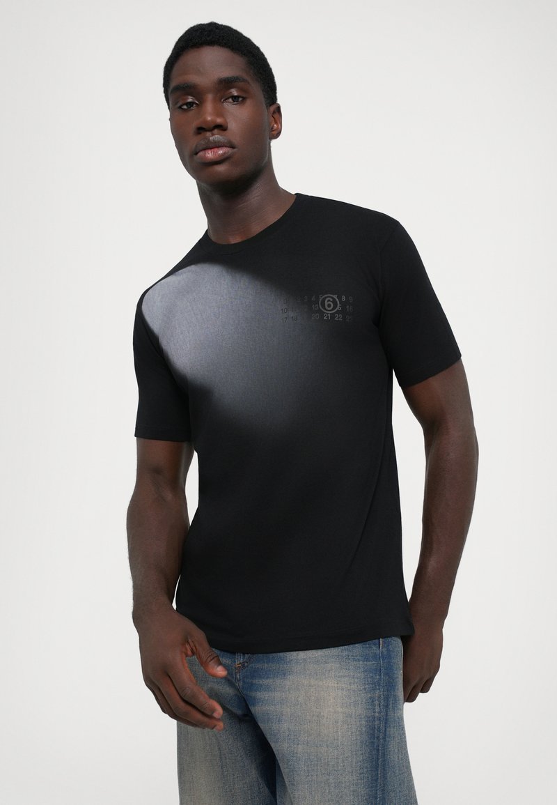 Sort T-shirt med korte ærmer, der har et gradient gråt mønster med et trykt design nær skulderen. Kombineret med lyseblå jeans.