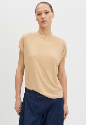 Frau trägt ein beigefarbenes kurzärmeliges Oberteil und dunkelblaue Hosen mit hoher Taille, steht vor einem schlichten, hellen Hintergrund.