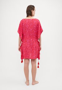 Donna in piedi a piedi nudi, che indossa un abito largo a fiori rossi e con motivo testuale, con frange, girata di spalle su uno sfondo bianco semplice.