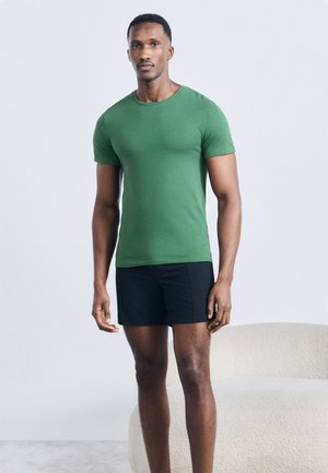 Homme debout portant une chemise verte à manches courtes et un short noir dans une pièce minimaliste avec un canapé de couleur claire.