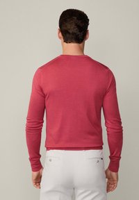 Roter, langärmliger Strickpullover mit Rundhalsausschnitt und gerippten Manschetten, von hinten gezeigt, kombiniert mit hellen Hosen.