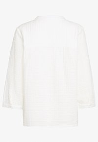 Camicia bianca a maniche lunghe con una vestibilità rilassata, polsini con bottoni e dettagli sul cucitura della spalla posteriore. Il tessuto ha una finitura stropicciata.
