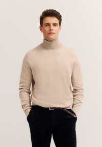Beige Rollkragenpullover mit langen Ärmeln, weicher Textur, taillierte Passform, kombiniert mit dunkelbraunen Cordhosen und einem geflochtenen Leder-Gürtel.