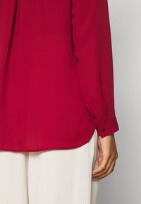 Blusa rossa con maniche lunghe, realizzata in tessuto leggero. Presenta polsini con bottoni e un orlo curvo sopra pantaloni bianchi texturizzati.