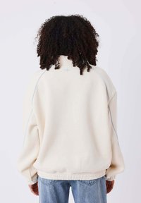 Felpa di colore crema con colletto alto, cuciture a contrasto blu, vestibilità rilassata e orlo a coste. Il tessuto sembra morbido con una texture liscia.