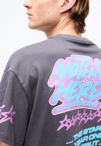 Άτομο με κοντά μαλλιά που φοράει σκούρο γκρι μπλουζάκι με φωτεινό μπλε και ροζ γραφικό κείμενο και αστέρια στην πλάτη και το μανίκι.