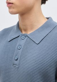 Νέος άνθρωπος που φοράει μπλε πλεκτό πόλο πόρο με τρία κουμπιά κουμπωμένα, με φαίνεται ο λαιμός και μερική προφίλ προσώπου.