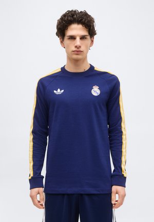 Νέος άνδρας που φοράει μπλε αθλητικό πουκάμισο με μακριά μανίκια και κίτρινες ρίγες, με λογότυπα της Ρεάλ Μαδρίτης και της Adidas, στέκεται μπροστά σε ένα απλό φόντο.