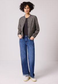 Donna con capelli ricci in piedi, indossa una giacca bomber con stampa animalier, top nero, jeans blu a gamba larga con risvolti e sneakers bianche.