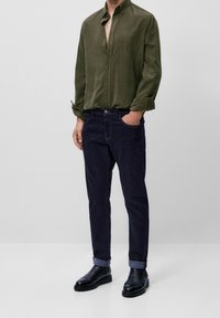 Homme portant une chemise olive débraillée, un jean noir roulé vers le haut et des bottes en cuir noires, se tenant avec une main dans sa poche.