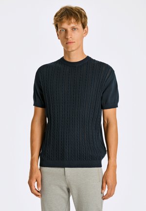Suéter de punto de manga corta en color azul marino con patrones de cable elevados; escote redondo y dobladillo acanalado; combinado con pantalones gris claro.