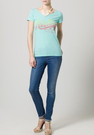 Camiseta de color verde claro con un escote de pico, que presenta un gráfico juguetón en amarillo y rojo. Combinada con jeans azules ajustados y sandalias de cuña color beige.