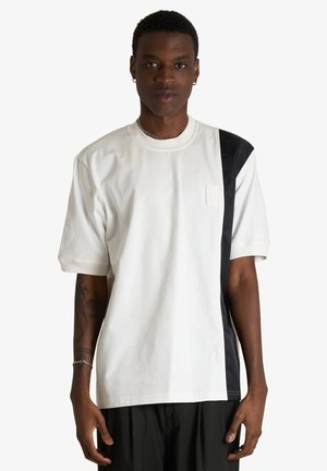 Giovane uomo con capelli corti che indossa una maglietta bianca oversize con una striscia verticale nera sul lato destro.