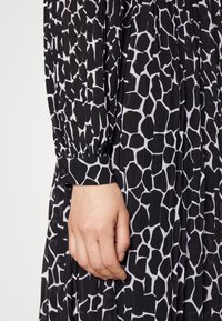 Χέρι γυναίκας resting δίπλα, φορώντας φόρεμα μαύρου και λευκού με σχέδιο καμηλοπάρδαλης και κουμπωτές μανσέτες.