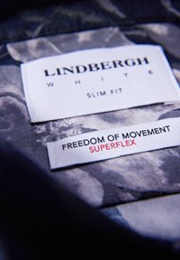 Etykieta slim-fit koszuli z białego materiału z napisami "LINDENBERGH WHITE" oraz "FREEDOM OF MOVEMENT SUPERFLEX" na wzorzystym niebieskim tle.