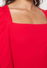 Czerwona rypsowa bluzka z kwadratowym dekoltem i bufkami, podkreślająca gładką teksturę oraz dopasowany krój.