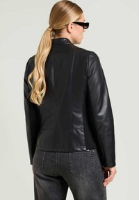 Vrouw met een lange blonde paardenstaart, gekleed in een zwarte leren jas, donkergrijze jeans en zwarte zonnebril, met de rug naar de camera gericht tegen een lichte achtergrond.