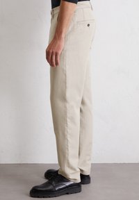 Beige cotton trousers with a straight fit, two back pockets with button closure, and worn with black lace-up shoes. Smooth texture.
