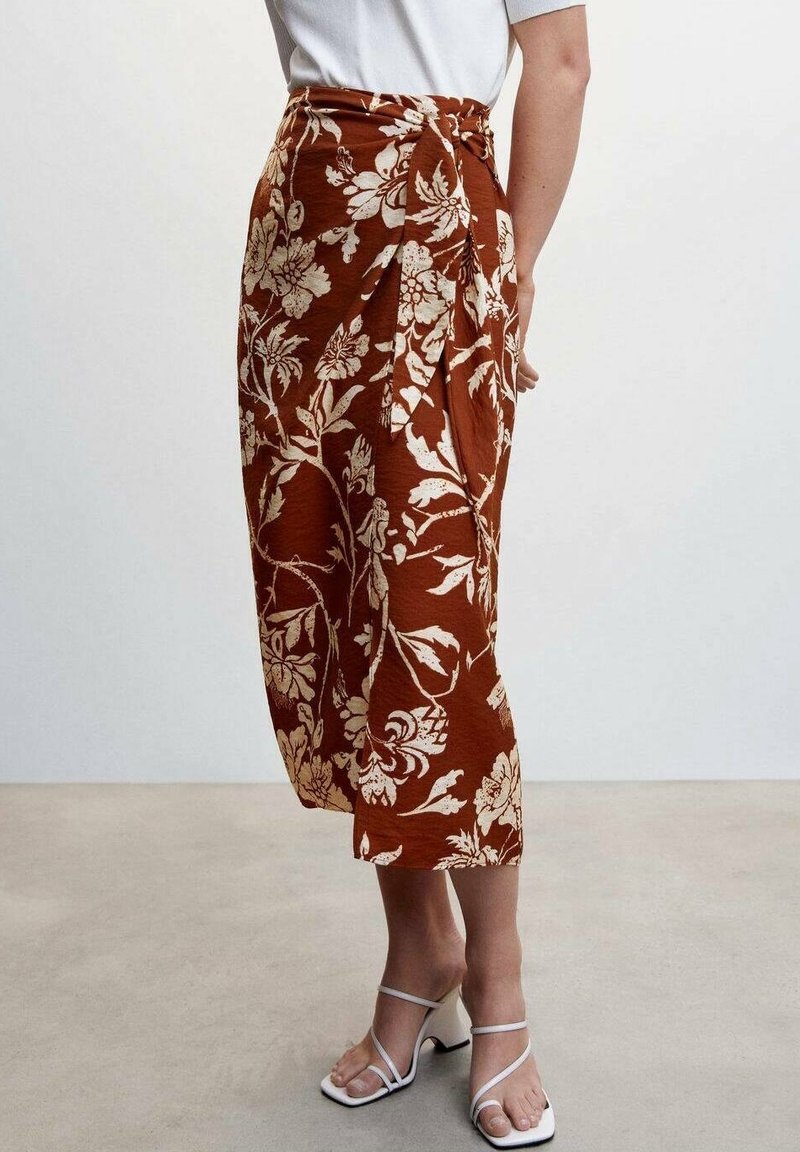Femme portant une jupe midi couleur rouille avec un imprimé floral blanc et des sandales blanches à talons avec brides, debout devant un fond uni.