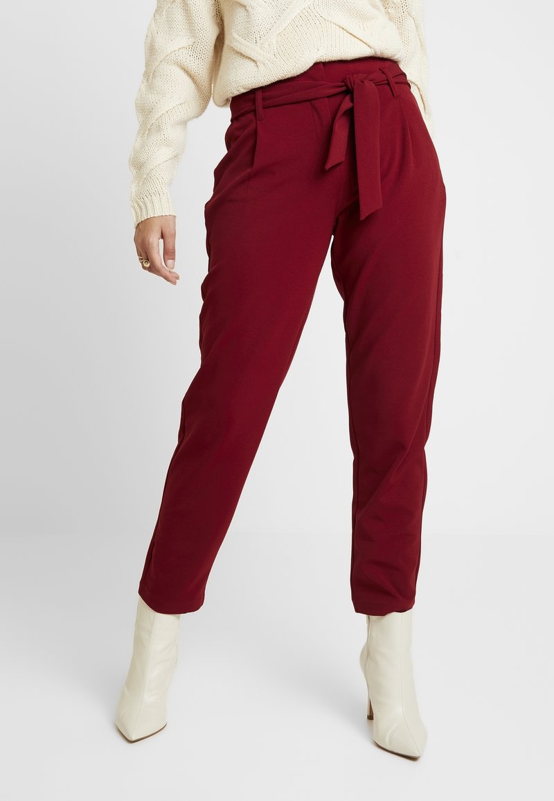 Pantalons cintrés bordeaux avec une taille haute ceinturée, présentant une texture lisse et une coupe fuselée, complétés par des bottines crèmes à talons aiguilles.