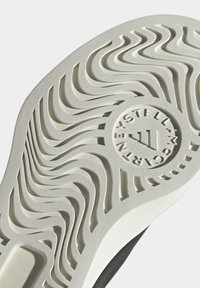 Semelle d'une basket blanche avec un motif de bande de roulement ondulé et un logo circulaire comportant le texte « STELLA McCARTNEY » et un dessin triangulaire au centre.