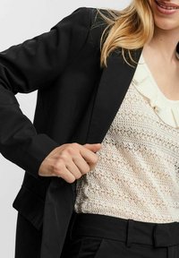 Blazer nero su una maglietta crema chiara e testurizzata con scollatura arricciata. La mano della modella sta aggiustando il rever del blazer.