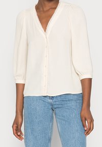 Cremefarvet bluse med V-hals, dekorativ blondekant og halværmer. Har knapper foran og blødt, glat stof. Parret med lyseblå jeans.