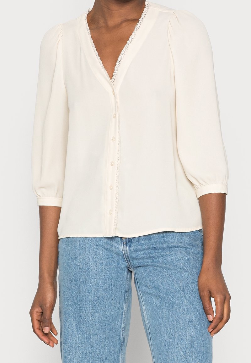 Cremefarvet bluse med V-hals, dekorativ blondekant og halværmer. Har knapper foran og blødt, glat stof. Parret med lyseblå jeans.
