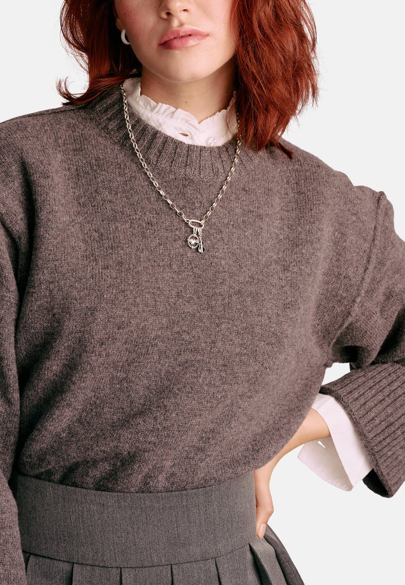 Brauner Strickpullover mit geripptem Ausschnitt, darüber ein weißes, gerafftes Hemd getragen. Eine silberne Kettenhalskette mit Anhängern ergänzt das Outfit.