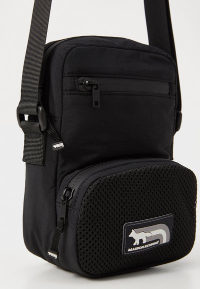 Sac bandoulière noir en tissu texturé, avec une poche zippée à l'avant, un accent en mesh, une bandoulière réglable et un patch de logo de la marque.