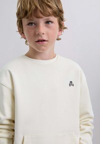 Crème-kleurige sweatshirt gemaakt van zachte stof, met een klein zwart doodshoofd en gekruiste botten embleem op de linkerborst.
