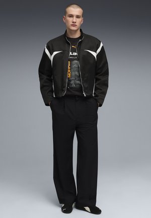 Mandlig model står med hænderne i lommerne iført sort-hvid lynlåstrøje, sort grafisk t-shirt, brede sorte bukser og sorte sneakers.