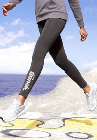 Grijze sportlegging met een aansluitend ontwerp, voorzien van witte logotekst op het linkerpand, gecombineerd met witte sneakers voor een sportieve uitstraling.