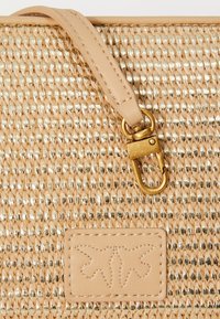 Superficie della borsa tessuta in beige e oro con tracolla in pelle marrone e patch con logo a farfalla cucito.