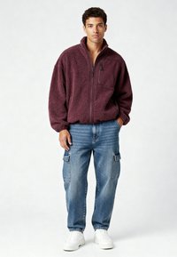 Giacca in pile bordeaux con zip frontale, tasche laterali e collo alto; abbinata a jeans cargo azzurri chiari e sneakers bianche.
