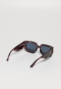 Hexagonal-frame sunglasses in dark tortoiseshell with dark lenses. Glossy finish, wide temples, and subtle branding detail on the side.