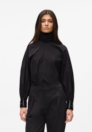 Femme aux longs cheveux foncés portant un chemisier noir à col montant avec des manches bouffantes et un pantalon noir assorti sur un fond uni.