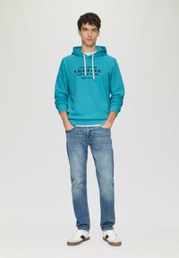 Türkise Kapuzenjacke mit Kordelzug, ausgestattet mit einem schwarzen Drucklogo, kombiniert mit hellblauen Jeans und weißen Sneakern mit schwarzen Akzenten.