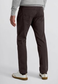 Homme portant un pantalon marron foncé et des baskets blanches, se tenant contre un fond clair et uni, vu de dos.