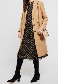 Camel wool coat over a black floral dress. Black knee-high socks and pointed ankle boots. Small handbag with brown and straw accents.