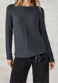 Femme portant un haut à manches longues texturé de couleur foncée avec une couture sur le devant, associé à un pantalon en velours côtelé foncé avec une taille nouée.