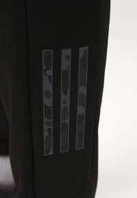 Pantalones deportivos negros con tres rayas verticales en un patrón de camuflaje, fabricados en un tejido suave y texturizado.