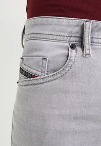 Grå denimjeans har en framficka med en liten röd detaljer och svart hårdvara. Tyget har en slät textur med synlig sömnad.